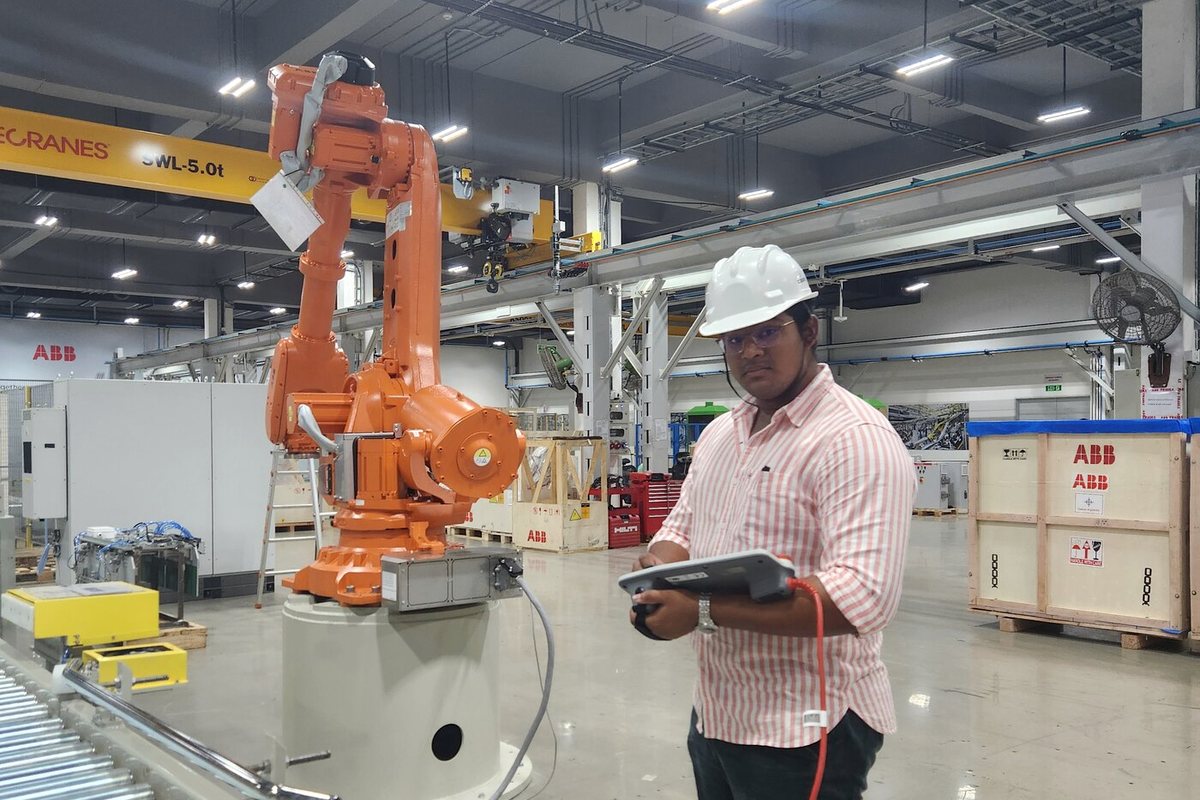A person in a white hard hat holding a teach pendant, standing beside a large orange ABB industrial robot on a factory floor with overhead cranes.
