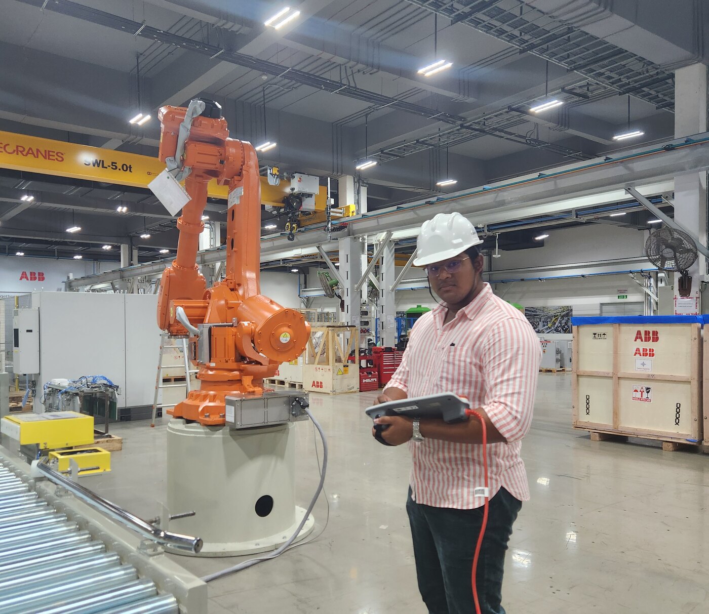A person in a white hard hat holding a teach pendant, standing beside a large orange ABB industrial robot on a factory floor with overhead cranes.