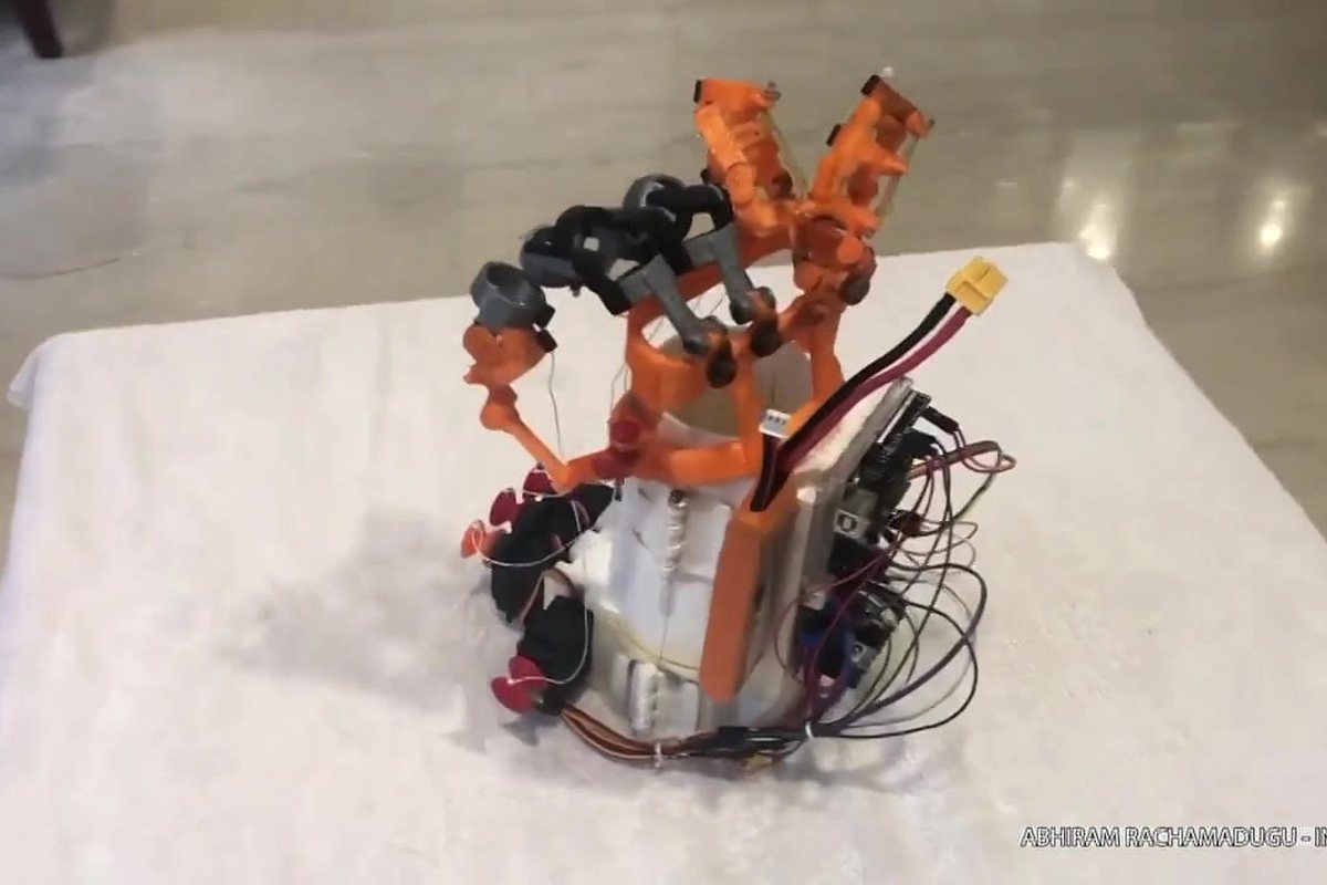 The bionic hand standing on a white cloth: orange printed fingers and tendon guides above a white forearm brace, with servos, wiring and a battery connector on the outside of the brace.