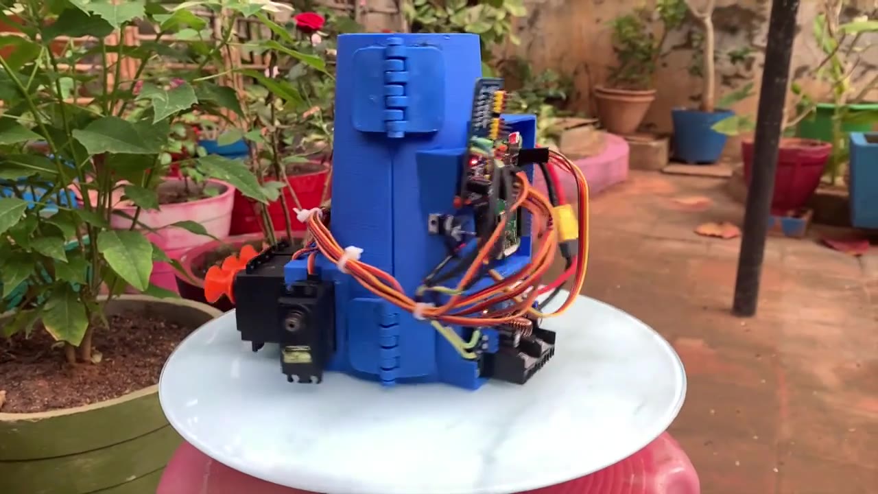 A blue printed forearm gauntlet on a small table in a garden, with servo mounts, a control board and a loom of orange and black wires.