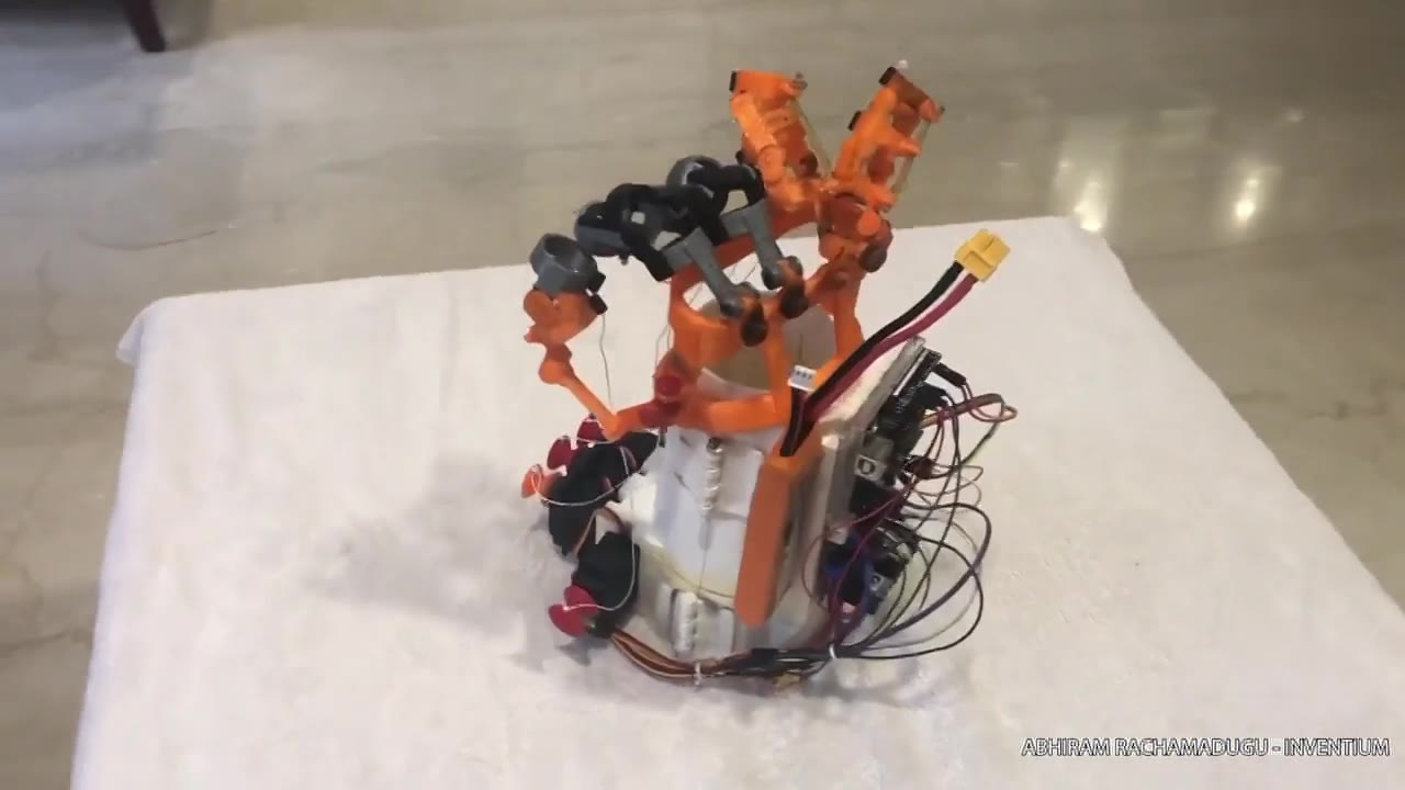 The bionic hand standing on a white cloth: orange printed fingers and tendon guides above a white forearm brace, with servos, wiring and a battery connector on the outside of the brace.