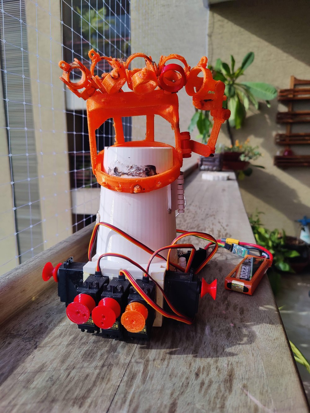 The printed arm standing on a wooden ledge outdoors: white brace, orange finger assembly, servo bank and a LiPo battery beside it.