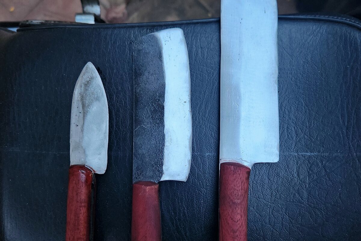 Three handmade kitchen knives with red wooden handles, blades polished, on a dark leather seat.