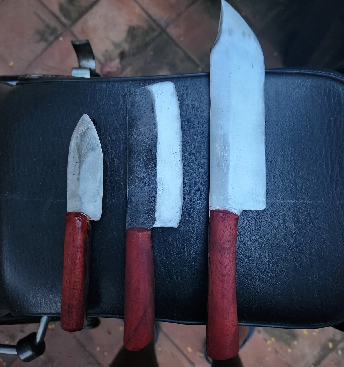 Three finished kitchen knives with red wooden handles laid side by side, blades polished, on a dark seat.