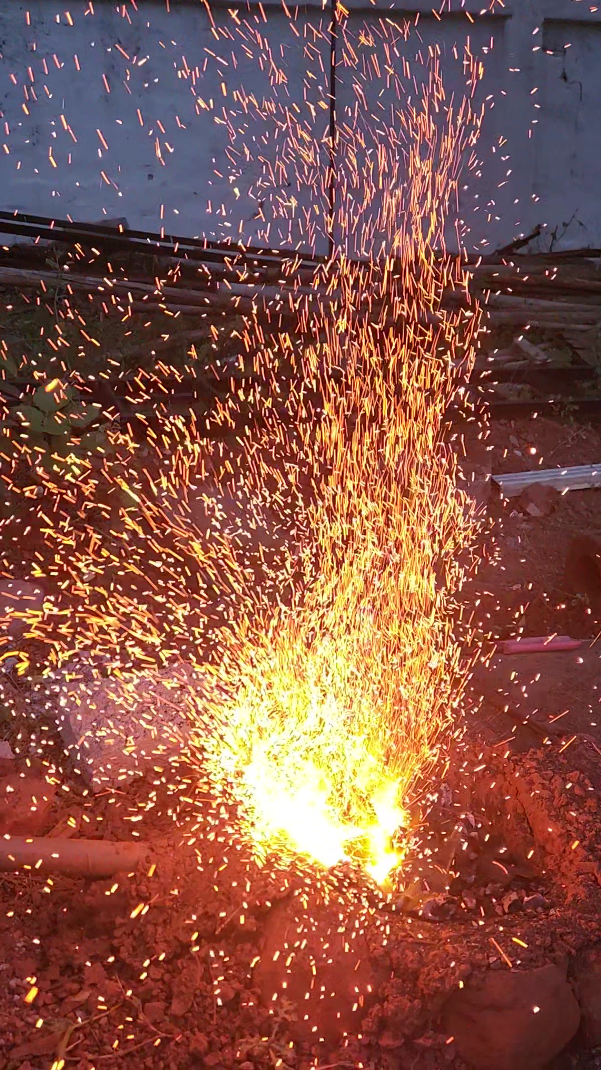 A charcoal forge at dusk throwing a tall fountain of orange sparks well above the fire.