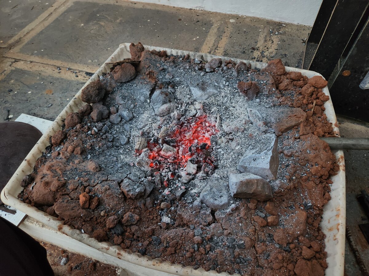 A raised forge hearth built into a plastered trough, lined with earth and stone, with charcoal glowing red in the middle.