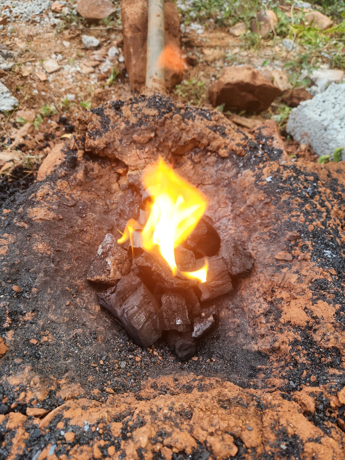 A shallow forge dug into red laterite soil, charcoal burning in the centre, a length of pipe entering from one side as a tuyere.