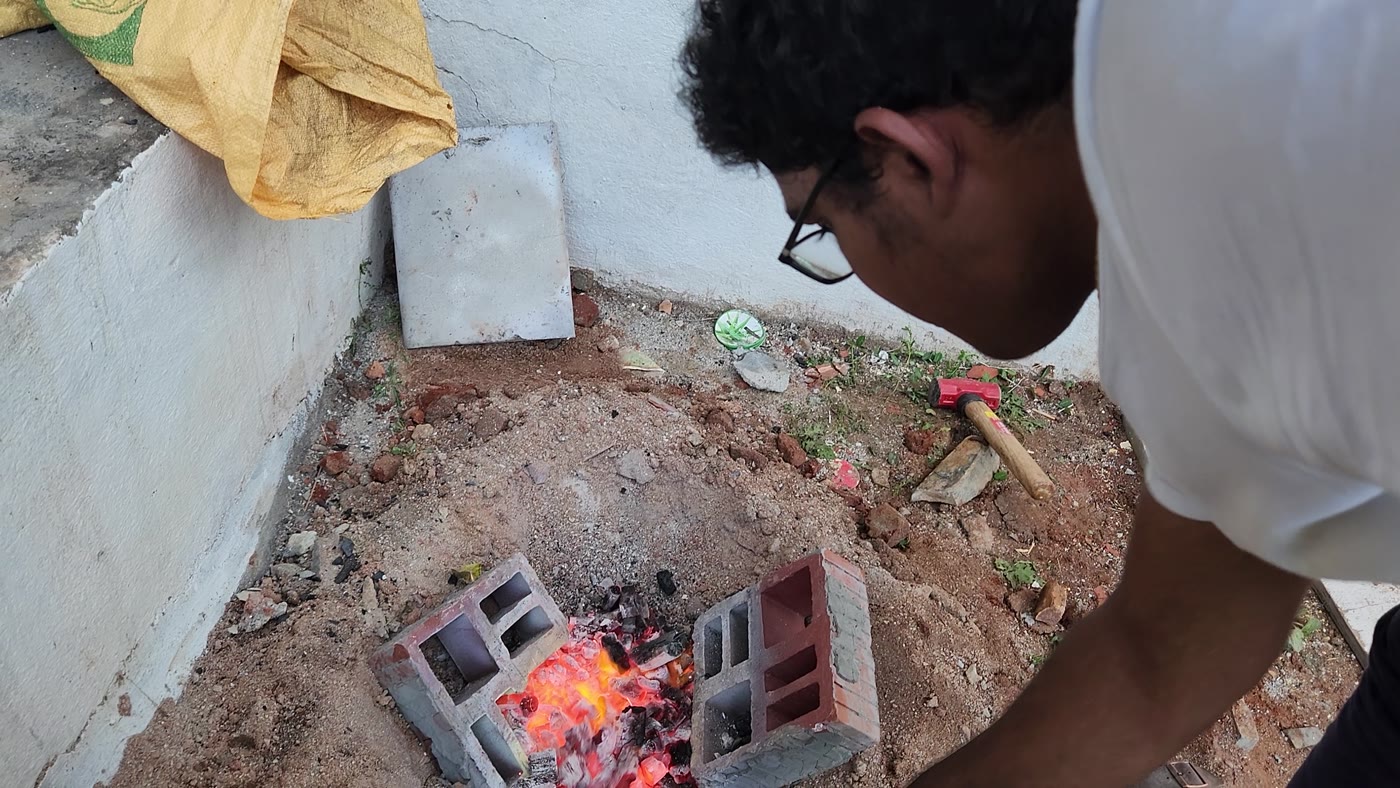 Working at a ground forge: a person lifting a stone slab beside a small block-walled hearth burning charcoal in a dug pit.