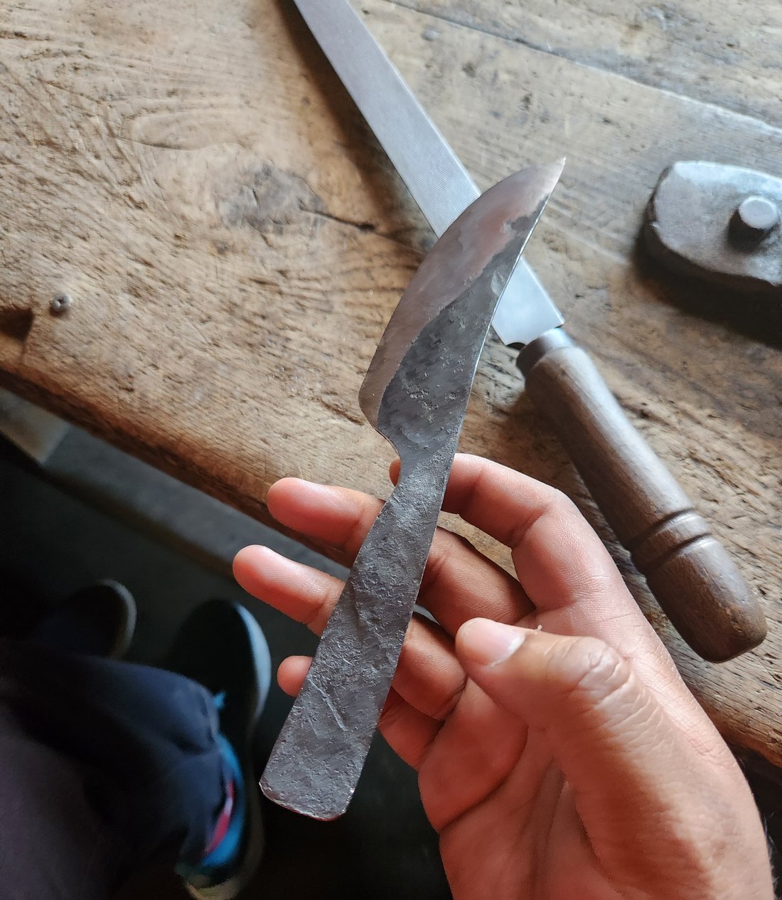 A rough-forged blade blank held in one hand over a worn timber bench, a wooden-handled file and a small stake tool lying behind it.