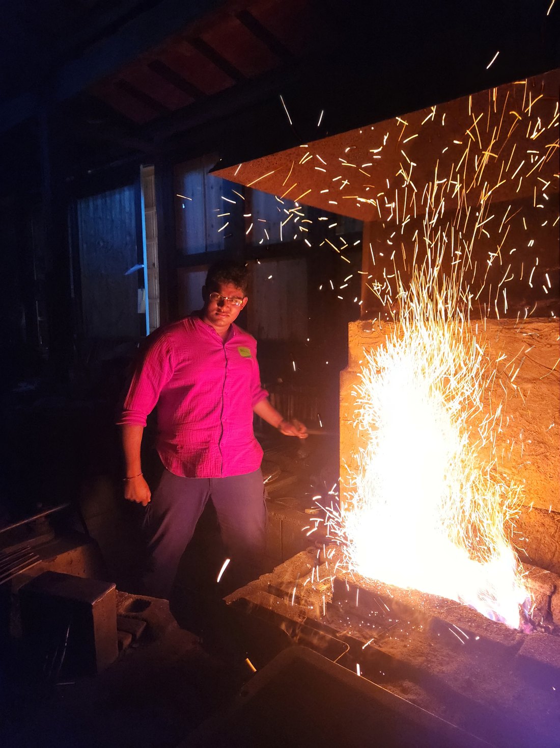 Abhiram at a forge in a timber-framed workshop, a dense column of orange sparks rising from the hearth beside him.