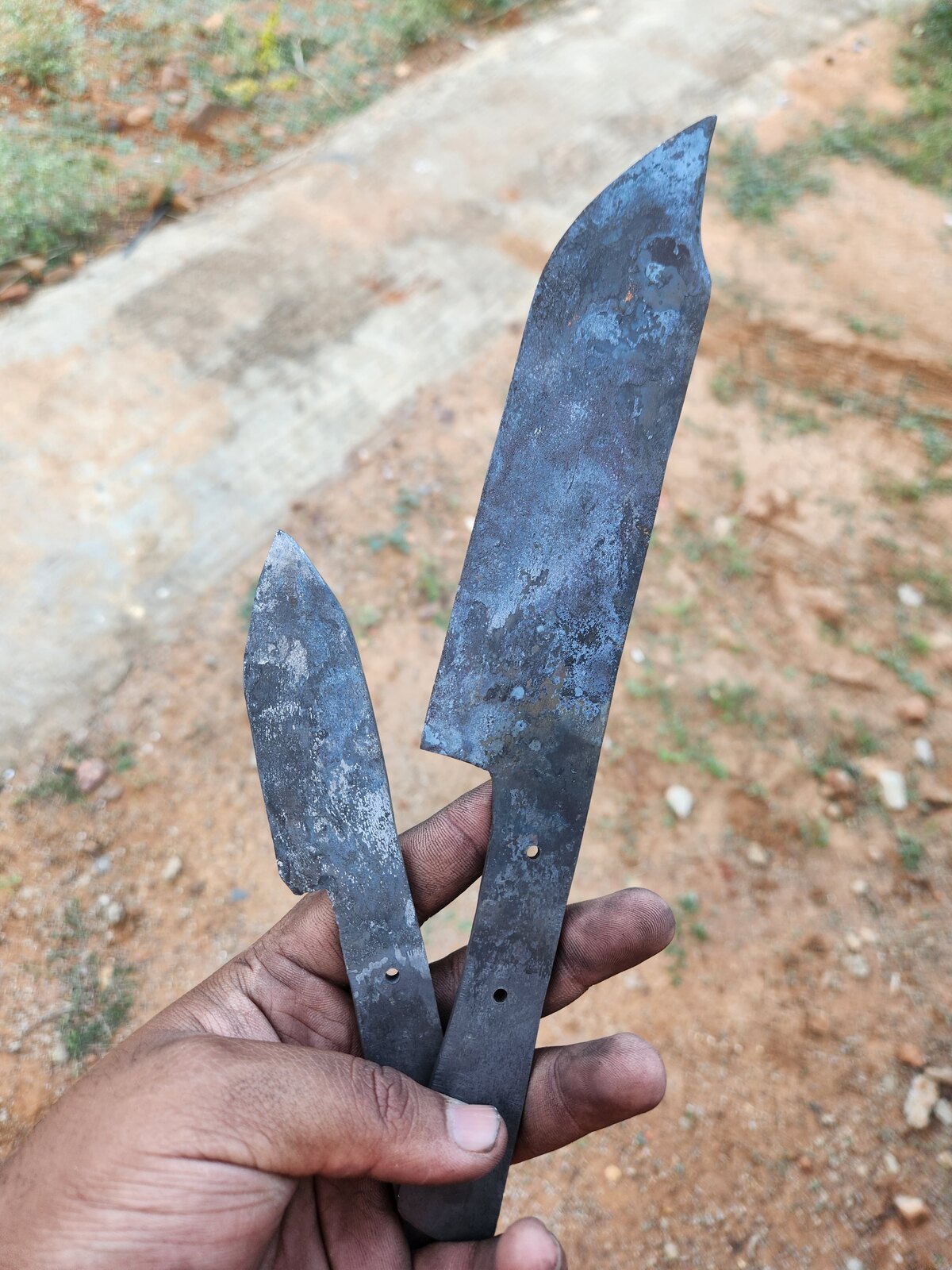 Two rough-ground blades held in one hand outdoors against red earth, still unhandled, drilled for pins.