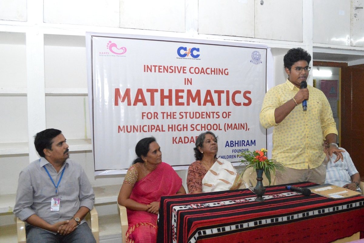 The maths coaching launch: Abhiram at a microphone and three school staff seated beneath the Children for Children banner.