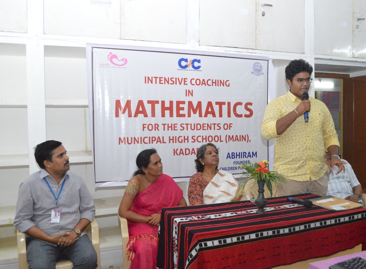 The launch event: Abhiram standing and speaking into a microphone at a table on a dais, three school staff seated beside him, beneath the programme banner.