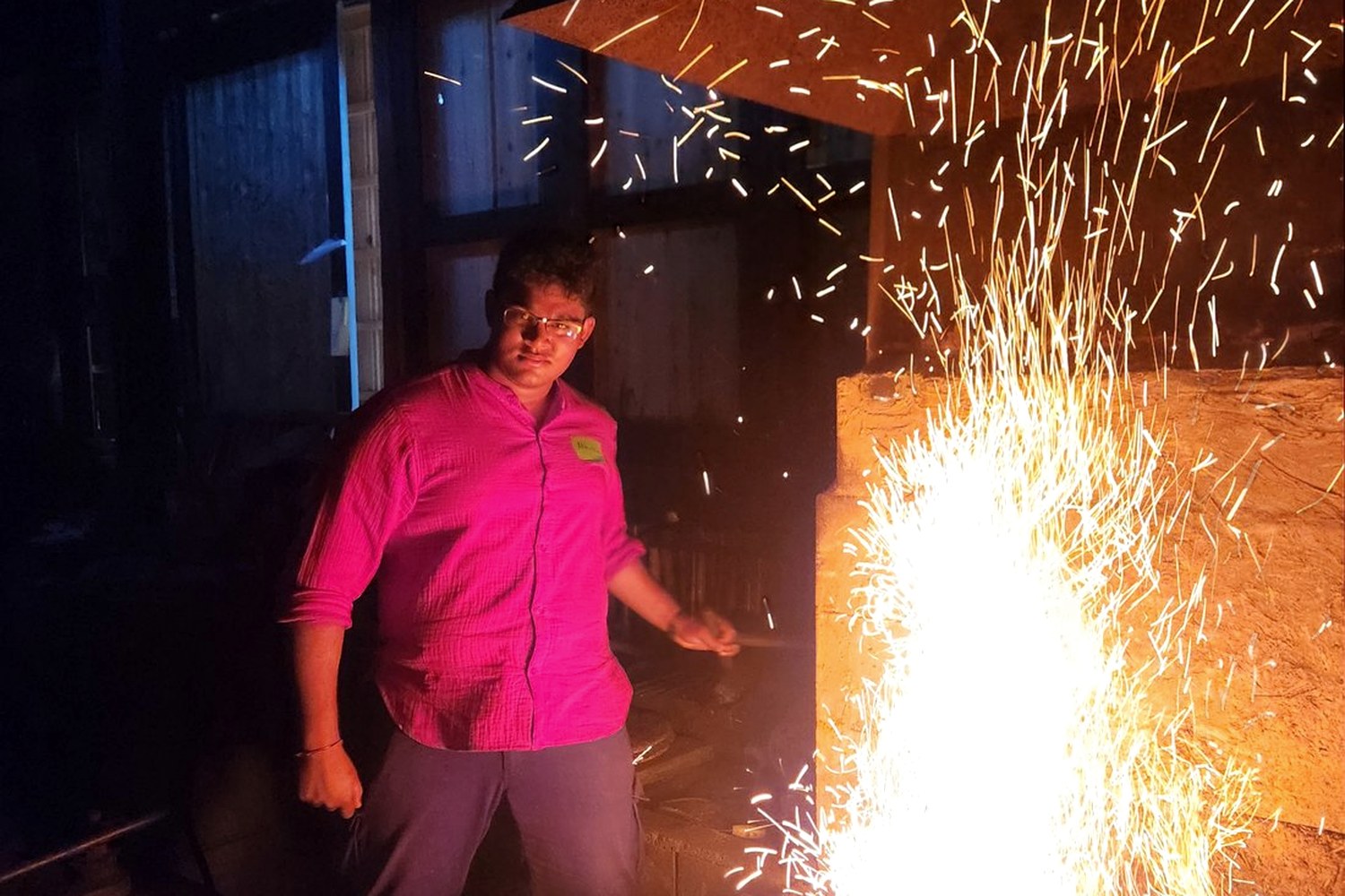 Abhiram at a forge in a master smith's workshop in Gifu, Japan, a fountain of orange sparks rising from the hearth beside him.