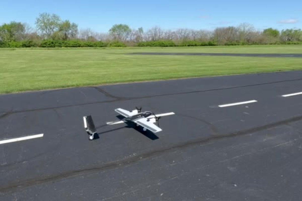 A twin-boom fixed-wing UAV lifting off from a paved runway, grass and trees beyond.