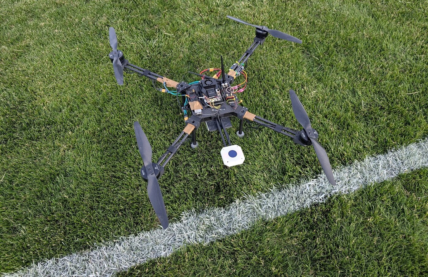 Skyfall v1, a carbon-fibre plate quadrotor with exposed wiring, sitting on a grass sports field beside a painted line.