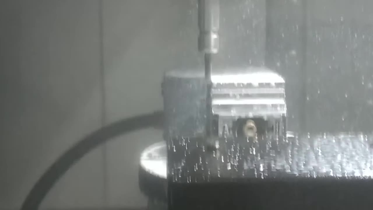 A 5-axis mill cutting an aluminium block held in a vice on a rotary trunnion table, coolant spraying off the end mill.