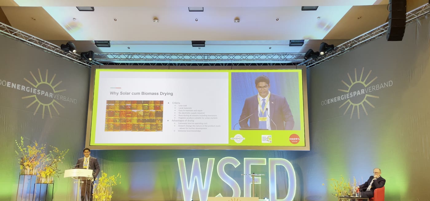 A conference stage lit green and blue with WSED lettering; a speaker at a lectern beside a screen showing a slide titled 'Why solar cum biomass drying'.